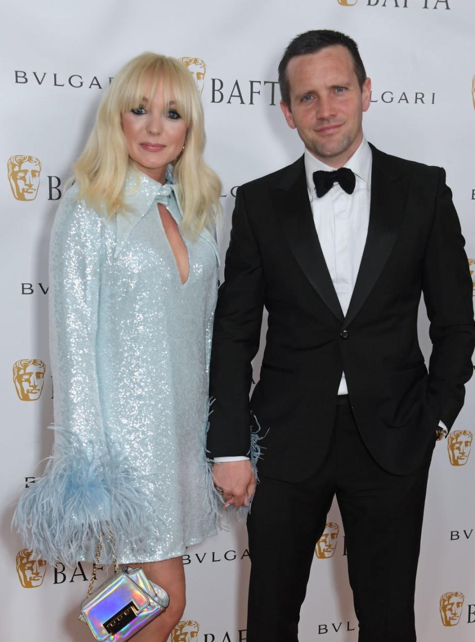 helen george and jack ashton stand holding hands and smiling at the camera, a woman with blond hair down to shoulders and fringe wears a silver sequin dress with feather sleeves, while her husband wears a black suit and bow tie