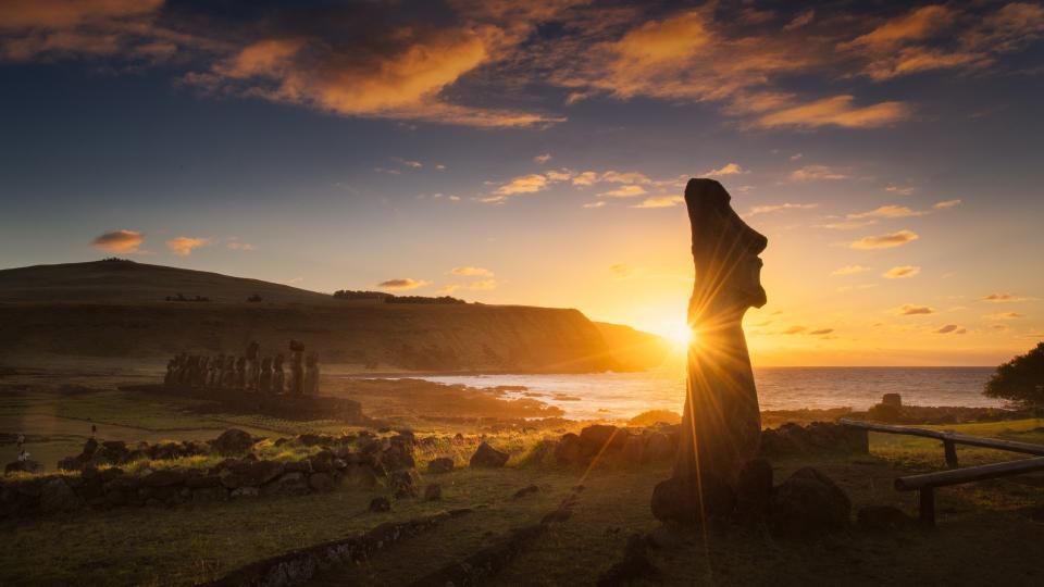  The sun sets behind a giant stone head on a lush green island. 