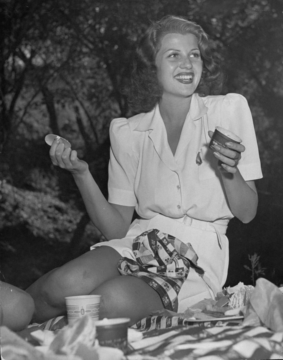 Actress Rita Hayworth, dressed in a white playsuit, sits on a picnic blanket in this undated photo.