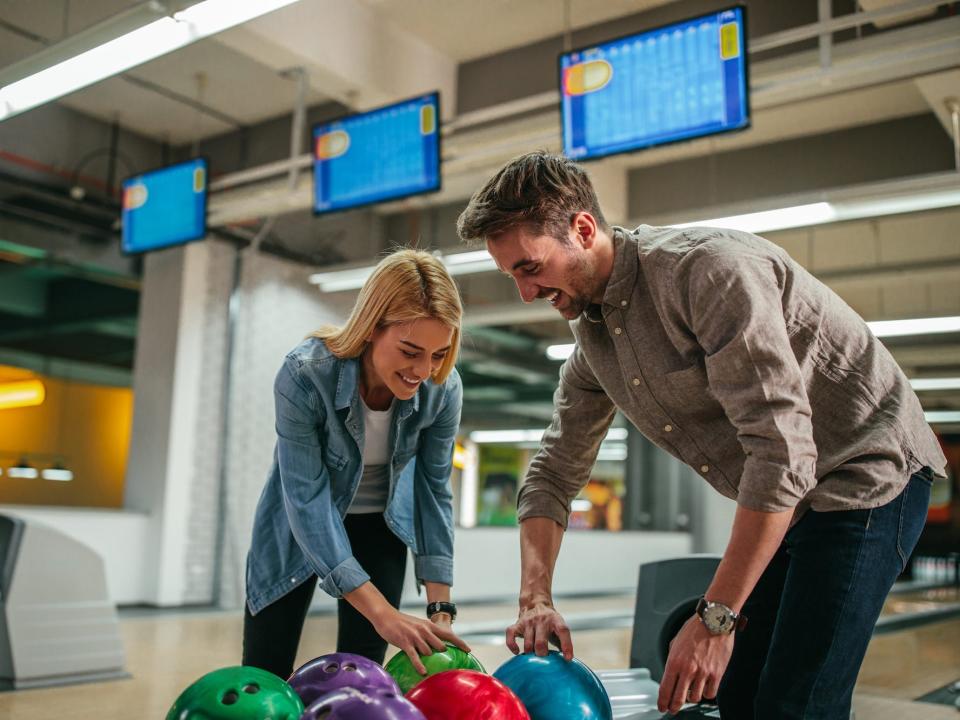 A woman and a man both reach for bowling balls.