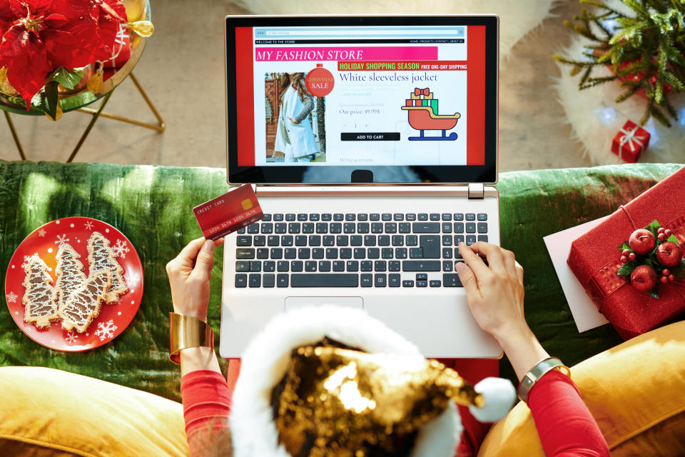 Holiday season. stylish female in sparkle gold sequin santa hat with credit card shopping clothes while using ecommerce site on a laptop while sitting on couch in the modern living room at Christmas.