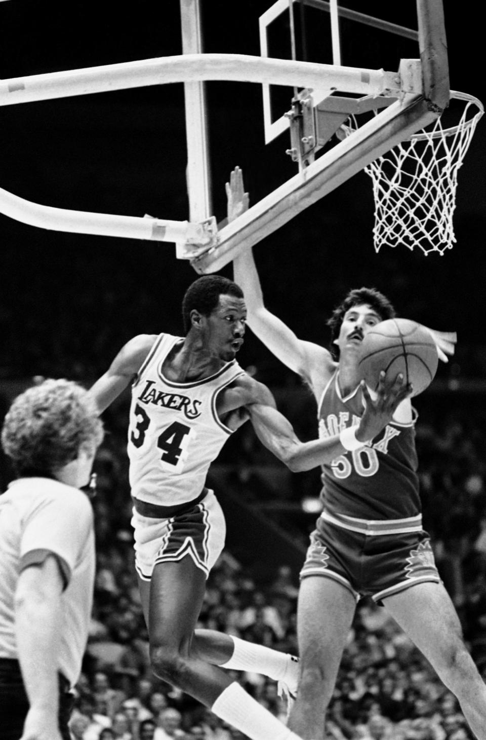 Clay Johnson (34) of the Los Angeles Lakers gives a little twist to the game as he goes up and out-of-bounds while trying to save the ball against the defense of Joel Kramer (50) of the Phoenix Suns at Los Angeles, April 28, 1982. The action came during the second half of Game 1 of the Western Conference playoff, which the Lakers won, 155-96.