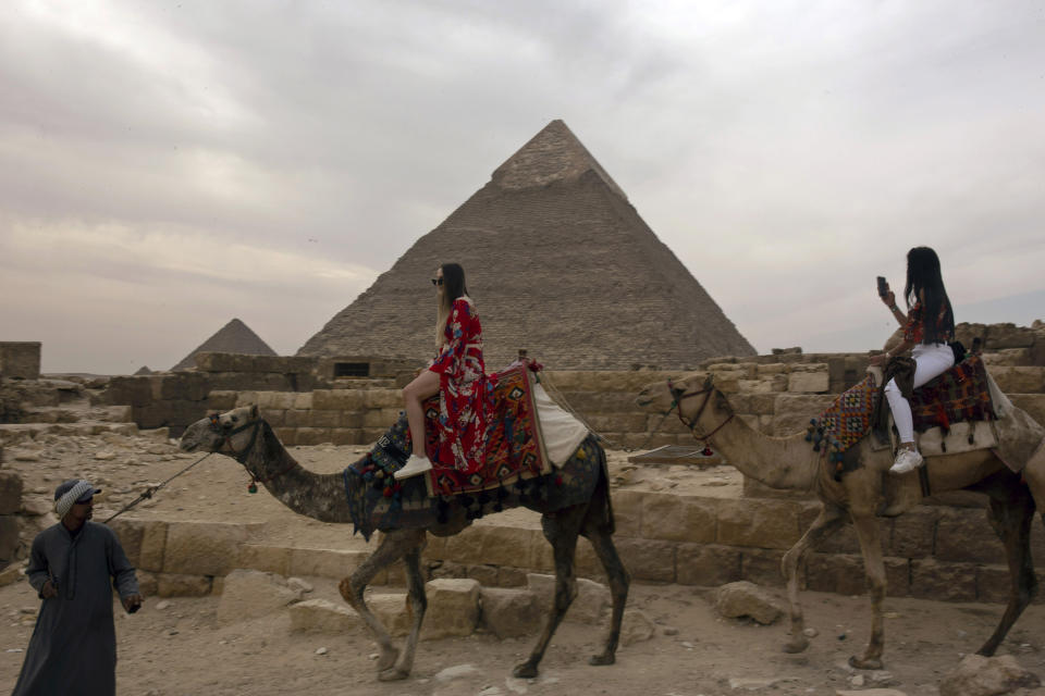 In this Tuesday, March 10, 2020, photo, two foreign women tourists ride camels at the Giza Pyramids near Cairo, Egypt. The country's prime minister announced at a news conference on Tuesday, March 24, 2020, a two-week, 7 p.m. to 6 a.m. curfew for its over 100 million people to slow the spread of the new coronavirus. The 11-hour curfew would go into effect Wednesday across the country and last for two weeks. (AP Photo/Maya Alleruzzo)