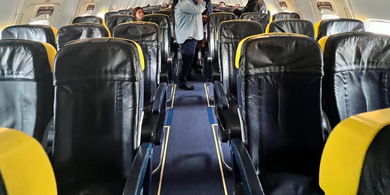 A Ryanair flight attendant helps a woman put her bag in the overhead bin while most of the seats remain empty during boarding, as viewed from the aisle.