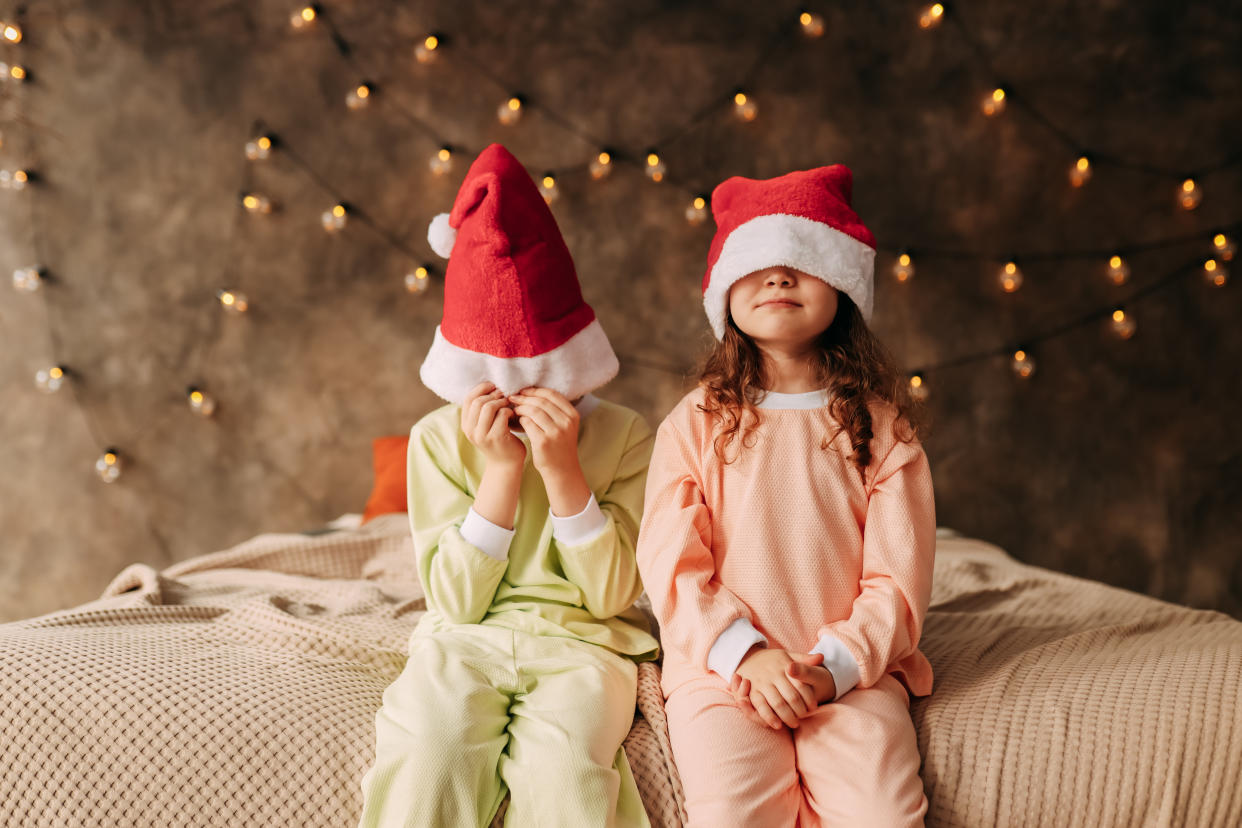 Cute funny little kids teenagers brother and sister twins in pajamas and Santa Claus hats have fun hugging and fooling around sitting on the bed in the cozy bedroom of the house during the Christmas holidays before sleep