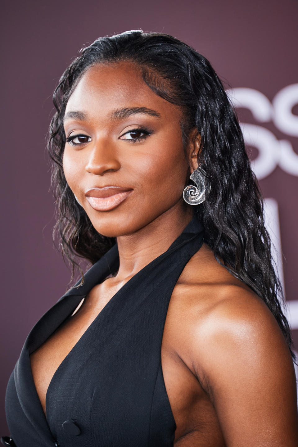 Normani on a red carpet, wearing a sleeveless black gown with a deep neckline, and spiral earrings