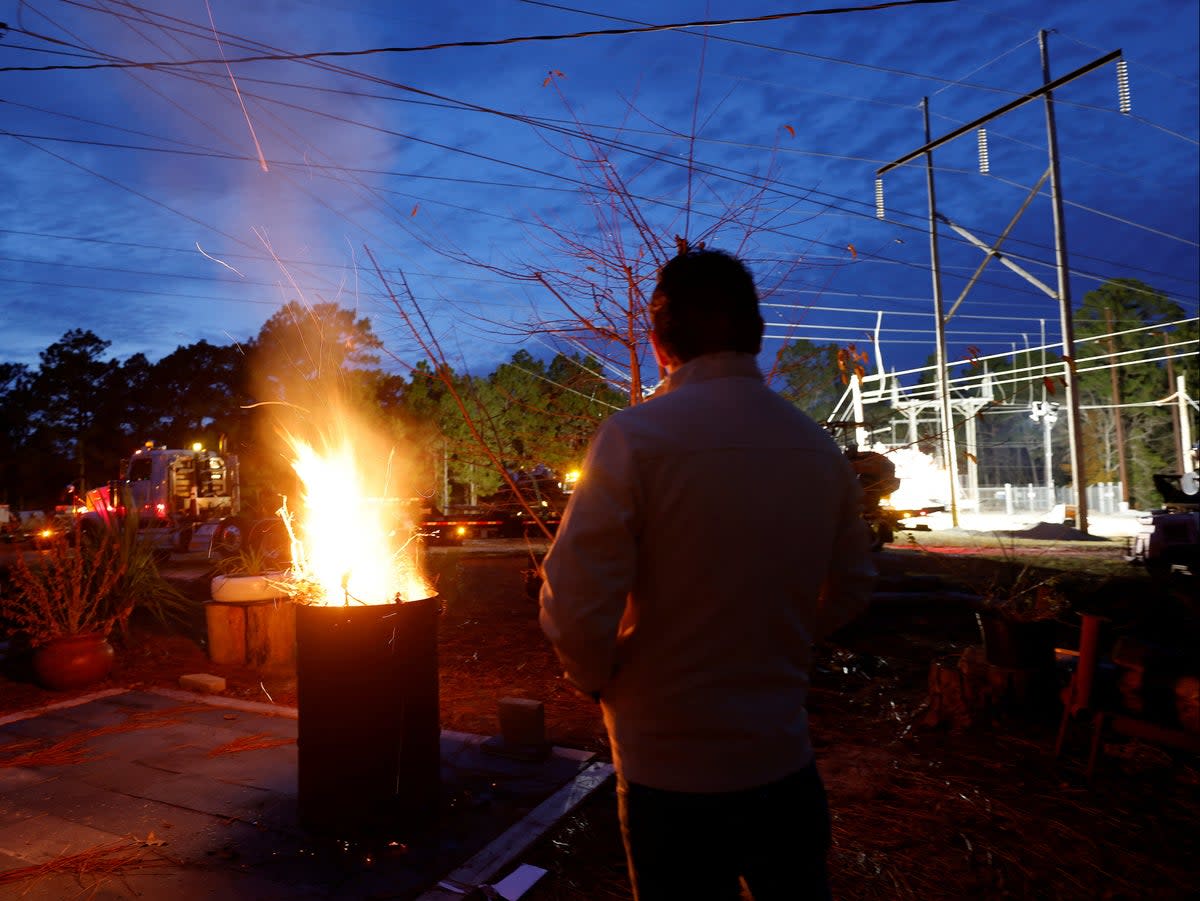 A man warms himself in front of a makeshift fire as he watches Duke Energy personnel work to restore power at a crippled electrical substation in Moore County, North Carolina (Jonathan Drake/Reuters)