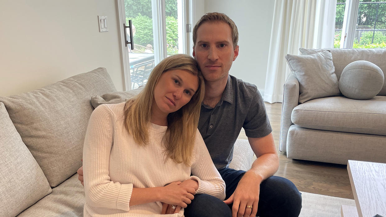 Chris and Brittany Wroblewski sit together on a couch near a sliding glass door and windows through which bushes and trees are visible.