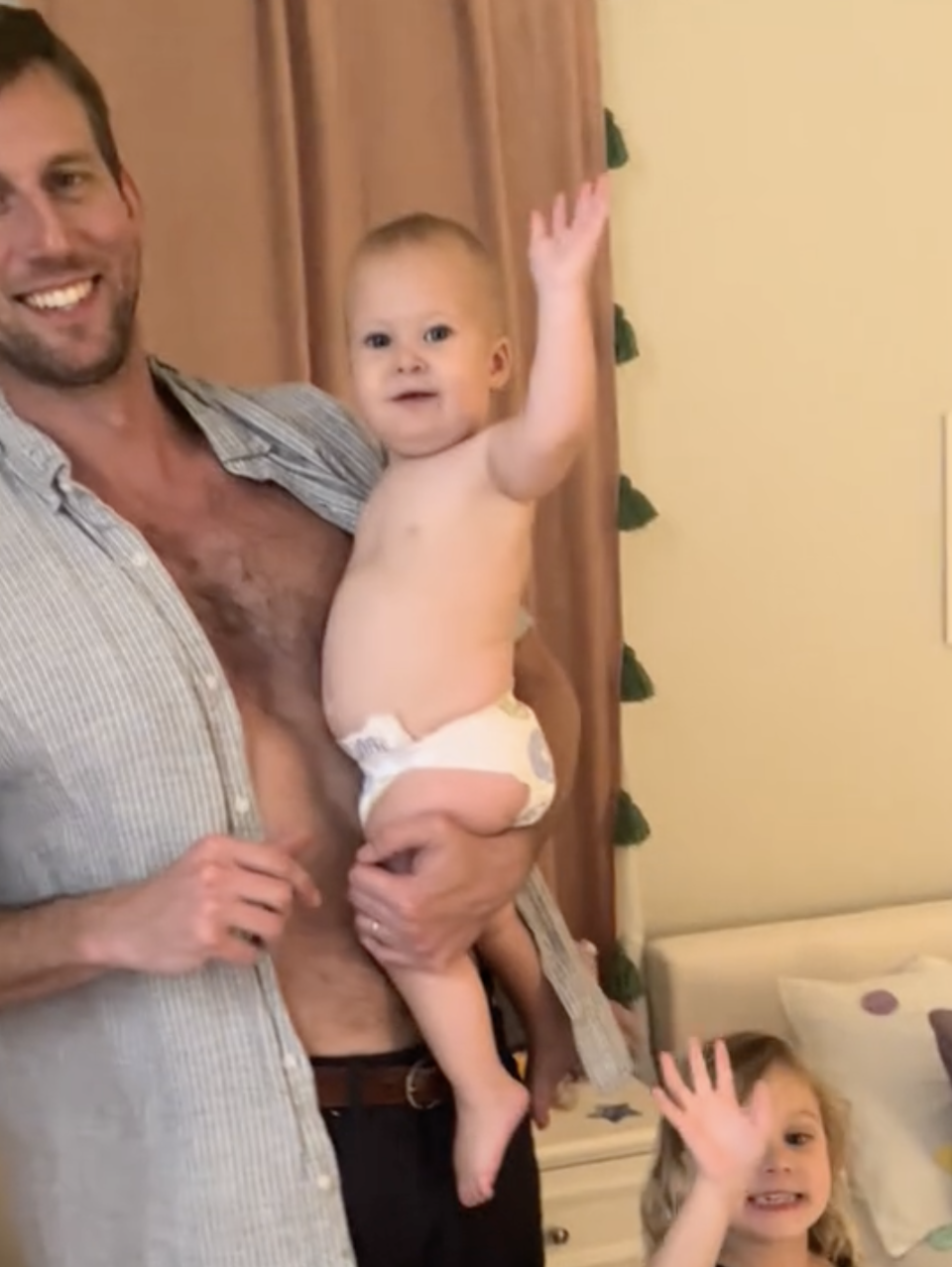 Man holding a baby wearing a diaper, with a young girl beside them, all smiling and waving at the camera