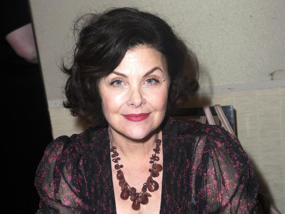 sherilyn fenn sitting at a table with her arms resting on the top, looking towards the camera and smiling