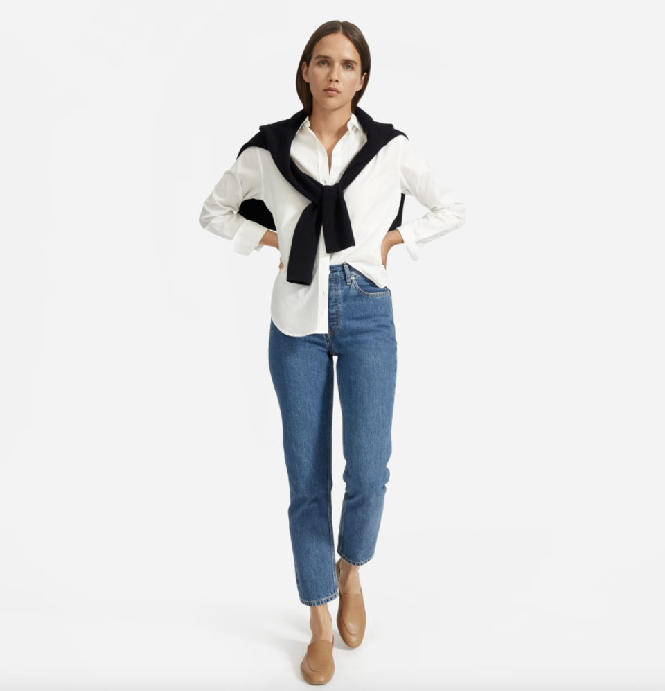 brunette model wearing blue jeans, black sweater, and white The Silky Cotton Relaxed Shirt in Off-White (Photo via Everlane)