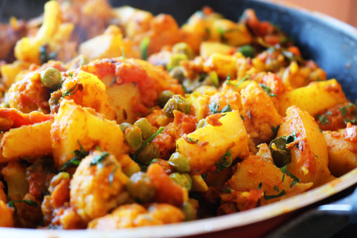 A close-up of a pan filled with a colorful vegetable curry, including pieces of potato, cauliflower, peas, and garnished with herbs