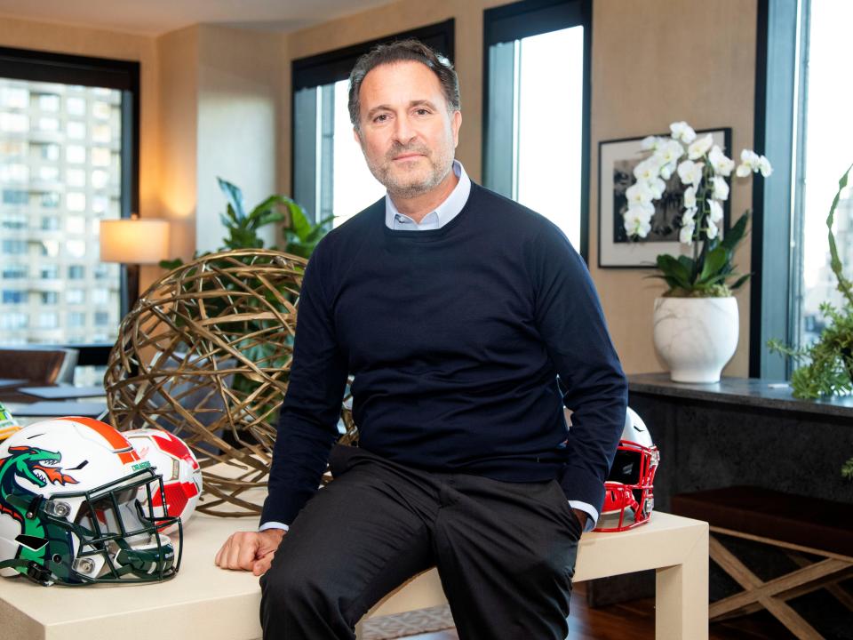 A photo of Gerry Cardinale, wearing a blue button down shirt, a navy sweater, and grey pants, leaning against a table in RedBird's conference room that is decorated with football helmets.