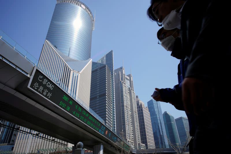 Pedestrians wearing face masks walk near an overpass with an electronic board showing stock information in Shanghai