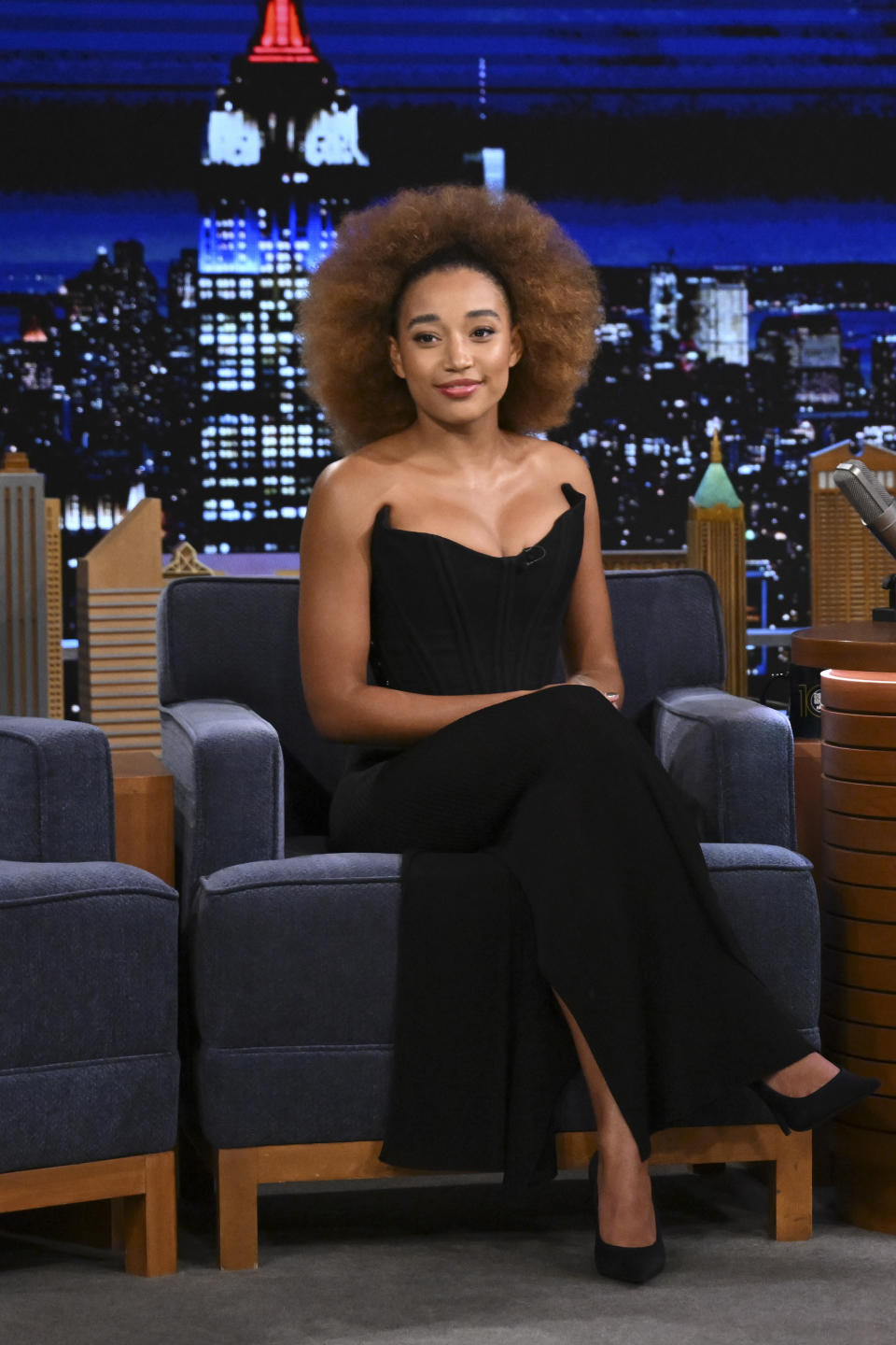 Amandla Stenberg sits on a talk show set in a sleek black strapless gown with a high slit, against a city skyline backdrop