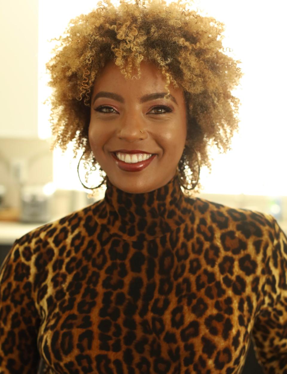 A woman with a blonde afro and a leopard shirt smiles to the camera