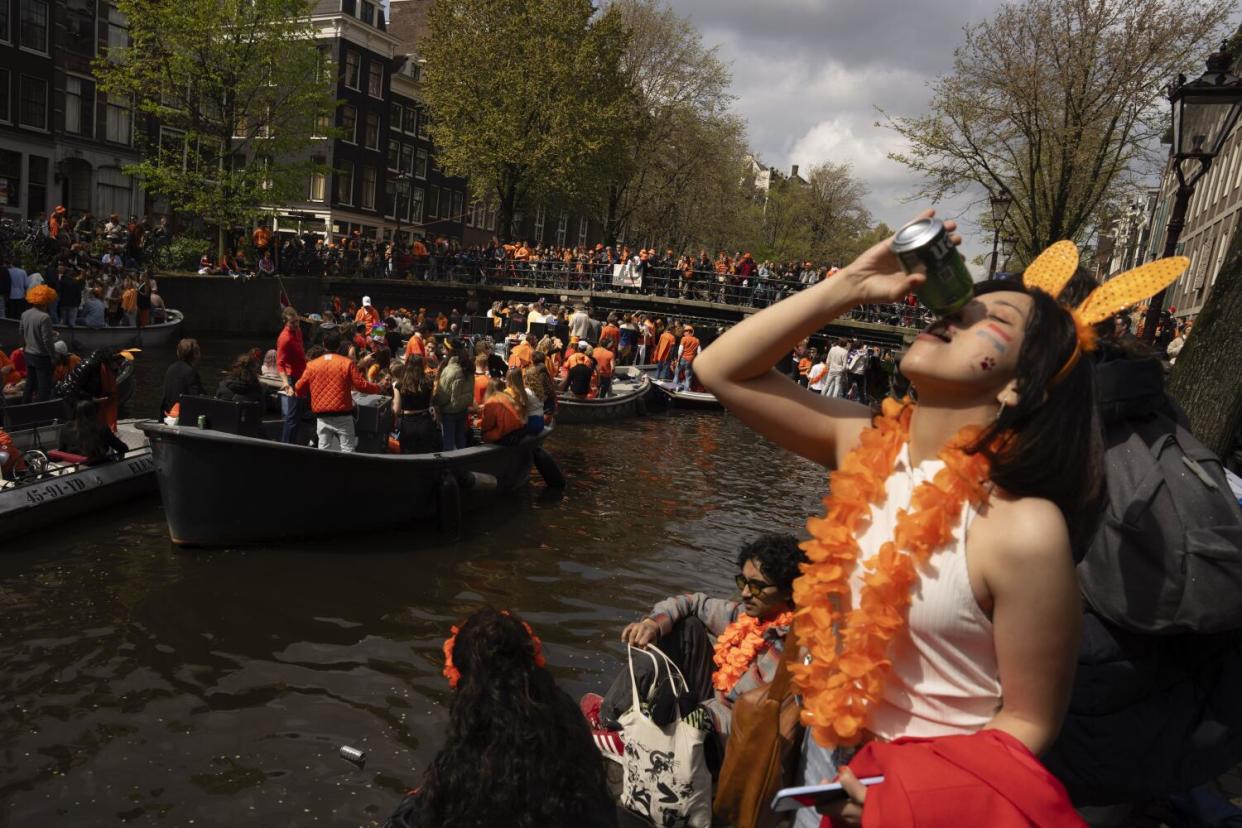 Boats full of people jam a waterway near a bridge crowded with pedestrians, as a woman in the foreground downs a canned drink