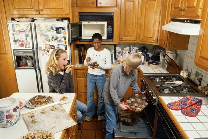 Teens baking in a kitchen, with one putting muffins in the oven and the others eating. The fridge is adorned with photos and magnets