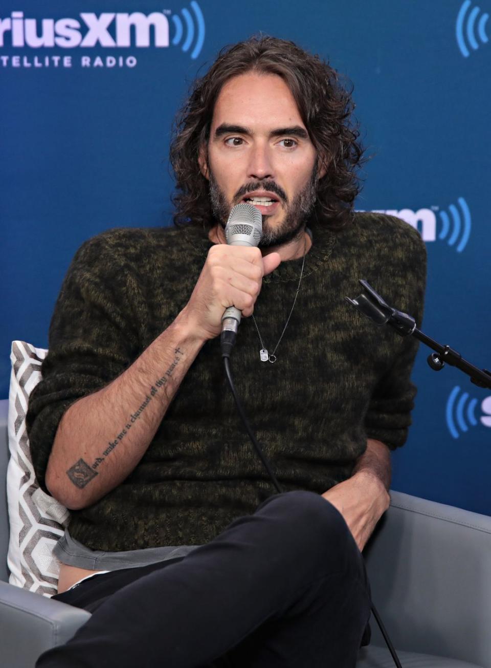 russell brand at an event in 2017, he sits in front of a blue background, talking into a microphone and wearing a dark jumper