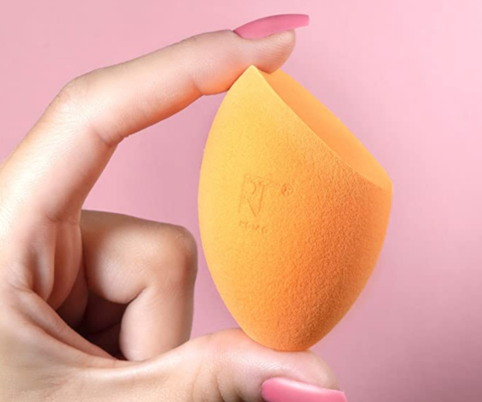 A close up of caucasian woman's fingers holding an orange beauty sponge against a pale pink background.