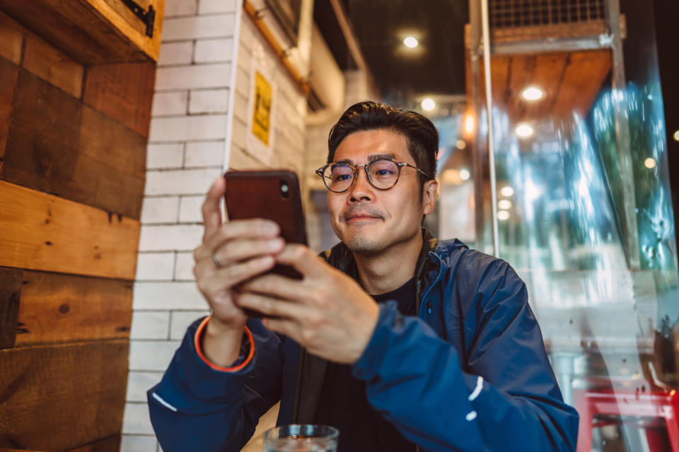 Man with glasses using a smartphone at a cafe, potentially exploring dating apps or communicating with a partner