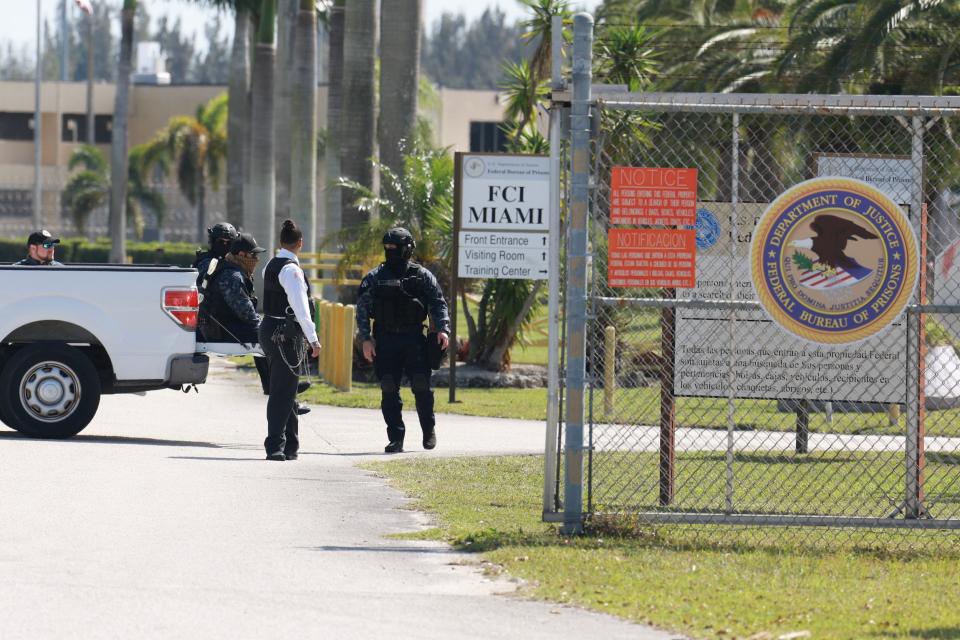 Security personnel stand guard near a wire fence. A white sign says FCI Miami