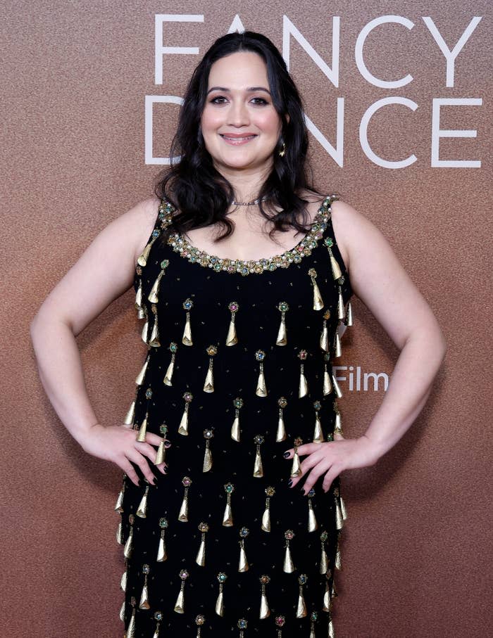 Unknown person on the red carpet with a sleeveless black dress adorned with decorative embellishments, posing in front of a "Fancy Dance" event backdrop