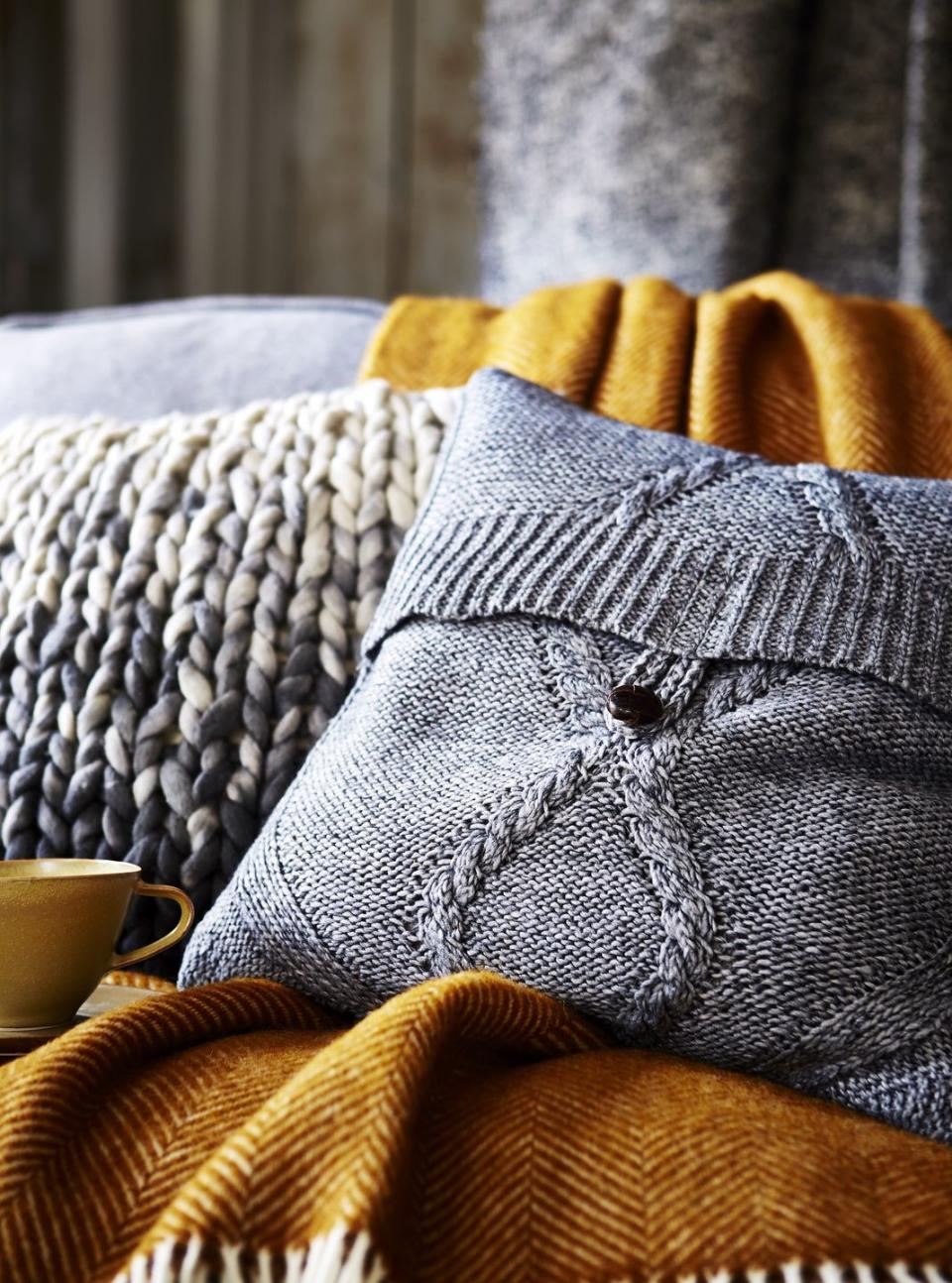 grey knitted woollen cushion cover with button fixing sitting on orange brown blanket, chunky knitted cushion cover behind, textured, soft, comfortable,