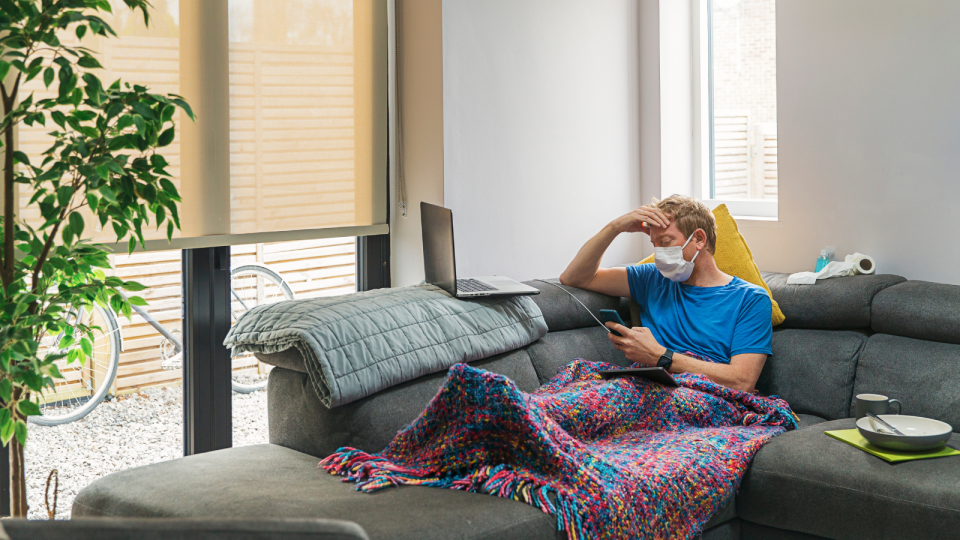 A man sits on a sofa looking upset while he wears a face mask and is covered by a blanket.