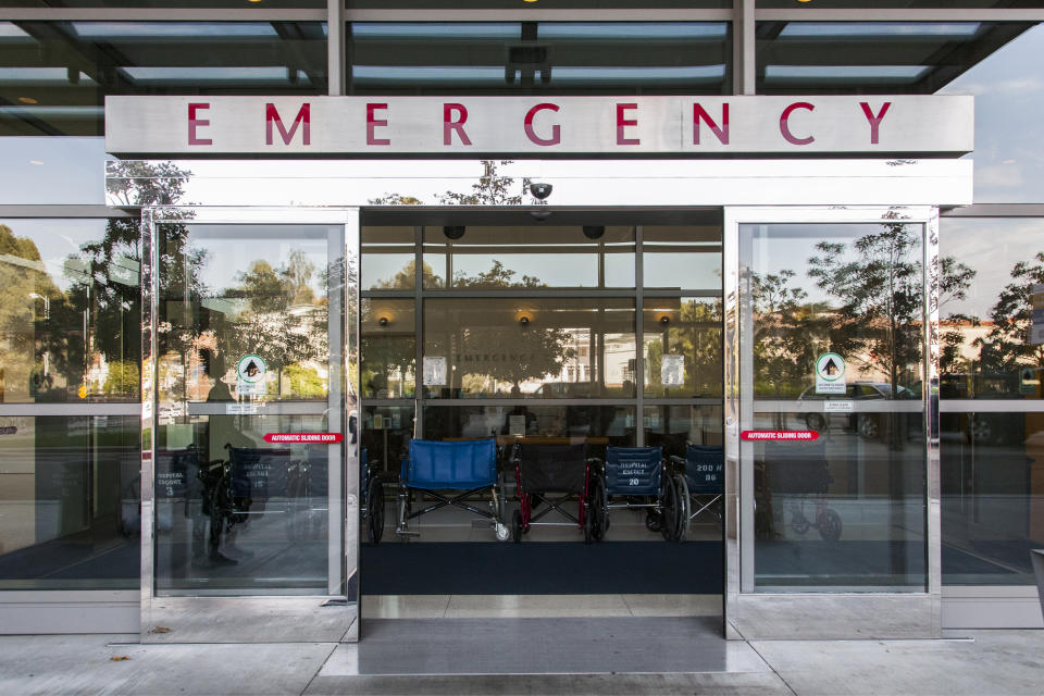 Entrance to an emergency room, with large glass doors, a sign above reading "EMERGENCY," and empty wheelchairs visible inside