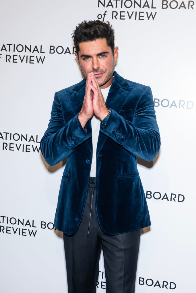 Zac Efron wearing a blue velvet jacket with hands together in a gesture, standing in front of a photo backdrop