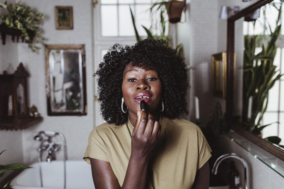 A woman is standing in a bathroom applying lipstick, looking into a mirror. She has curly hair and is wearing a casual outfit