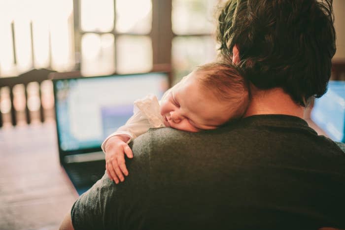 A parent holding a sleeping baby on their shoulder, both seated in front of a laptop