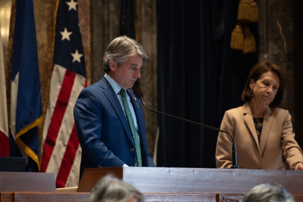 Sen. Henry stands at his desk with a microphone. He is wearing a blue suit and green tie.