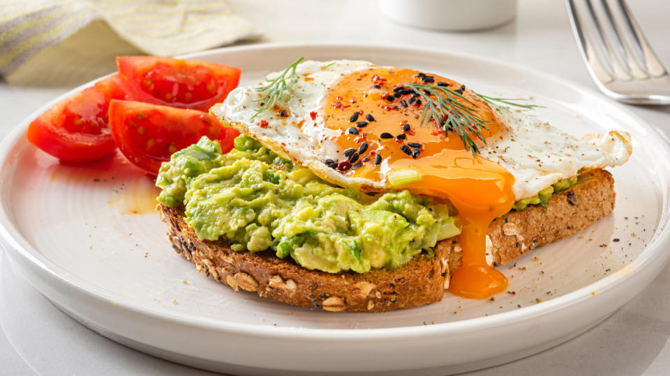 A white plate with toasted topped with mashed avocado, an over-easy egg and sliced tomatoes, which are some of the best cortisol-lowering foods