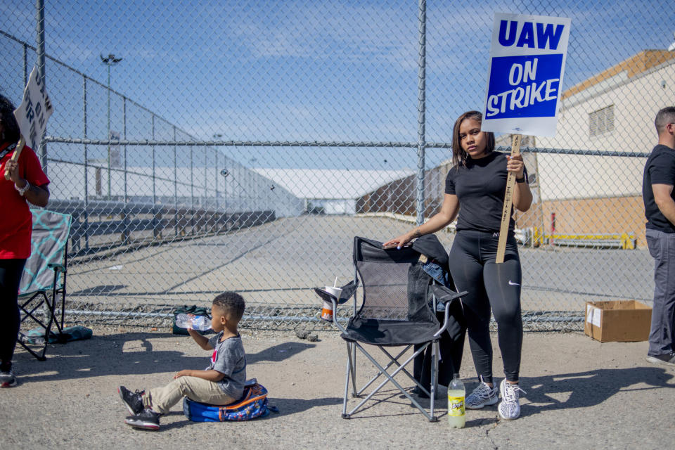 Rylan Hurt, 2 of Flint, drinks bottled water while sitting on his backpack, trying to stay cool as Flint resident Delani Richardson, who has worked as a temp on the trim line for six months, stands tall on the picket line outside of the Flint Assembly Plant during the fourth day of the national UAW strike against General Motors on Thursday, Sept. 19, 2019 in Flint, Mich. (Jake May/The Flint Journal via AP)