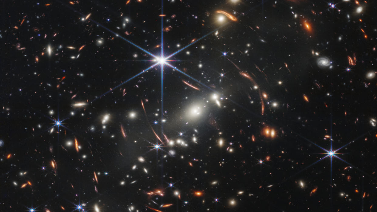  A view of space with lots of specks of light indicating galaxies and a few streaks representing lensed light. 