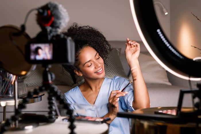 A young adult swatches different colors of eyeshadow on their arm as they record a makeup tutorial video at home.