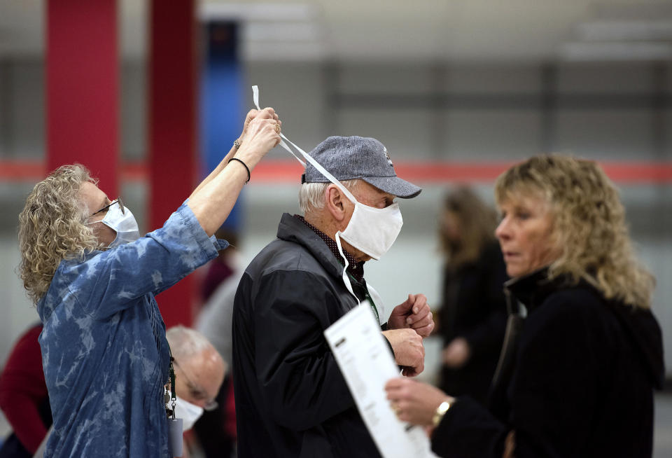 Poll worker Patty Piek-Groth, left, helps fellow poll worker Jerry Moore, center, put on a mask to prevent the spread of coronavirus, as the polls open for the presidential primary election at the Janesville Mall in Janesville, Wis., on Tuesday, April 7, 2020. Hundreds of voters in Wisconsin are waiting in line to cast ballots at polling places for the state's presidential primary election, ignoring a stay-at-home order over the coronavirus threat. (Angela Major/The Janesville Gazette via AP)