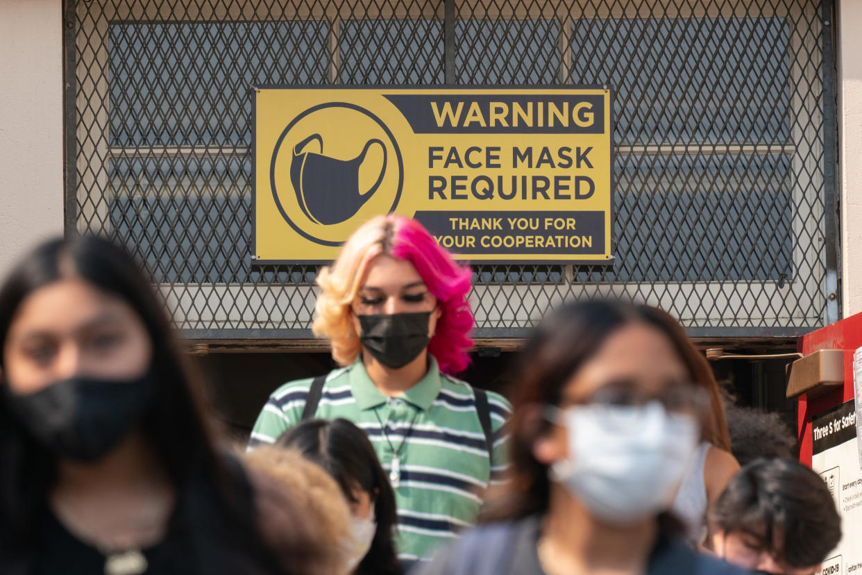 Students wearing face masks walk below a sign reading: Warning, face mask required. Thank you for your cooperation.