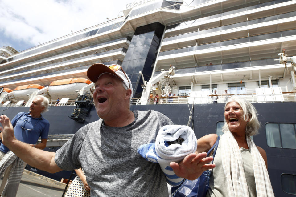 Passengers react after they disembarked from the MS Westerdam, back, at the port of Sihanoukville, Cambodia, Friday, Feb. 14, 2020. Hundreds of cruise ship passengers long stranded at sea by virus fears cheered as they finally disembarked Friday and were welcomed to Cambodia. (AP Photo/Heng Sinith)