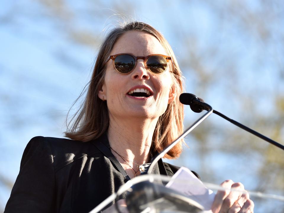 Jodie Foster speaks into a microphone outside, wearing sunglasses