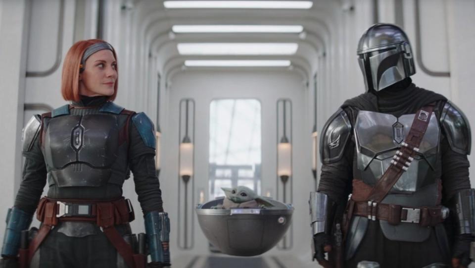 a helmetless Bo-Katan and Din Djarin look at each other while standing in a hallway with Grogu floating in his pod betwene them on The Mandalorian