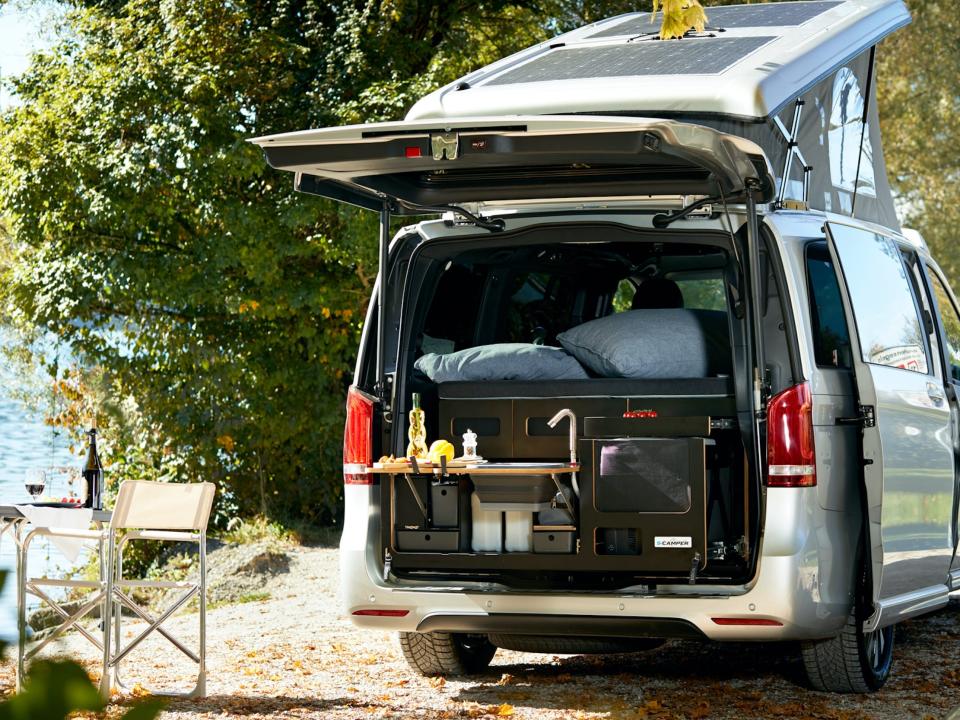 The trunk of the EQV open, revealing a mini kitchen. The van is parked next to an outdoor dining set.