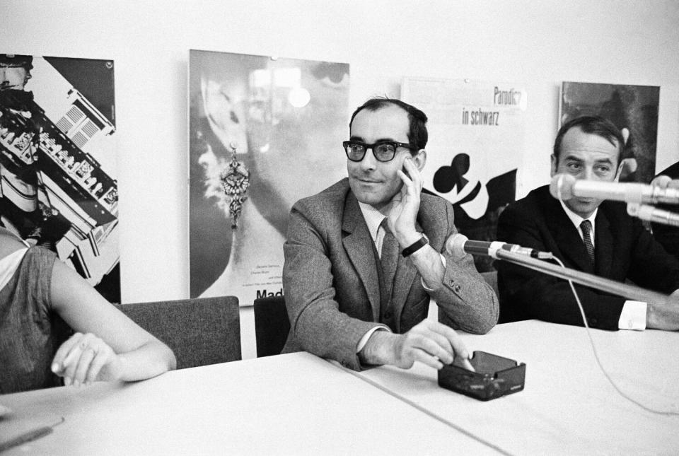 French director Jean-Luc Godard sits at a table with other people and microphones.