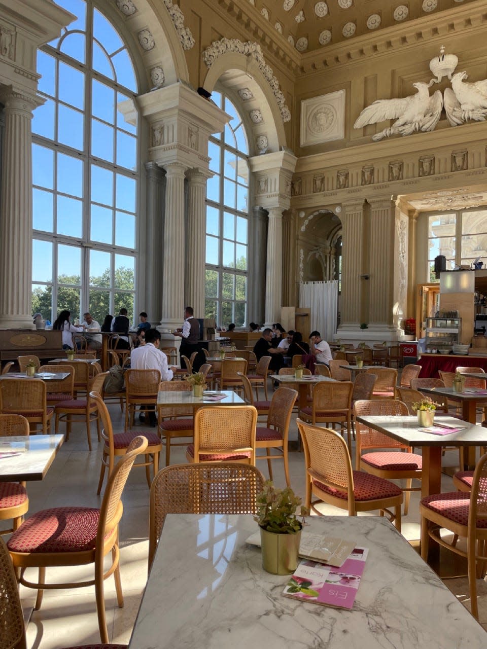 A cafe with columns and large windows and rattan chairs in front of marble tables in Vienna