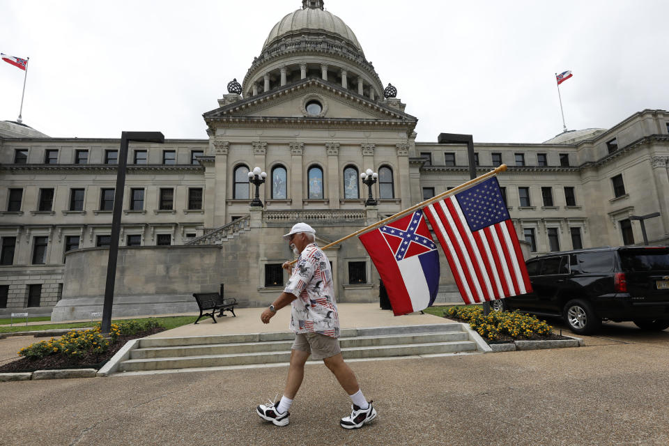 File-This June 27, 2020, file photo shows Don Hartness of Ellisville, Miss., walking around the Capitol carrying the current Mississippi state flag and the American flag in Jackson, Miss. Mississippi will surrender the Confederate battle emblem from its state flag, more than a century after white supremacist legislators embedded it there a generation after the South lost the Civil War. Mississippi's House and Senate voted in succession Sunday, June 28, 2020, to retire the flag, with broad bipartisan support. Republican Gov. Tate Reeves has said he will sign the bill, and the state flag would lose its official status as soon as he signs the measure. (AP Photo/Rogelio V. Solis, File)