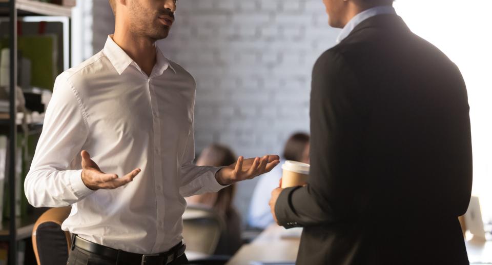 Two men having a conversation in an office setting. One gestures with open hands while the other holds a coffee cup. Their expressions are engaged