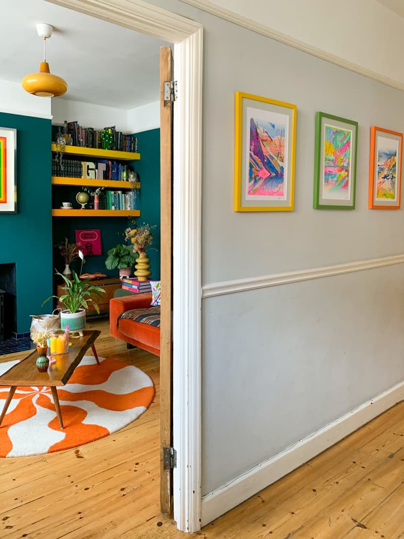 hallway, colorful art, colorful frames, wall molding, door frame, teal wall, orange and white wavey stripe circle rug, wood floors, coffee table, yellow rounded lighting fixture, yellow book shelf, orange couch, plants