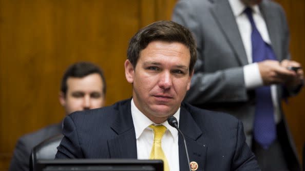 ron desantis stares at the camera while sitting in a wood paneled room, he has a display screen, microphone and name plate in front of him, he wears a black suit jacket, white collared shirt and yellow tie